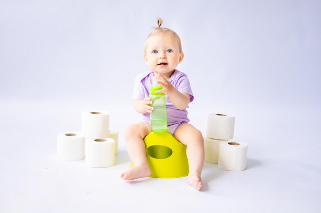 Une jolie petite fille souriante est assise sur un pot avec une bouteille d'eau et des rouleaux de papier toilette isolé sur fond blanc