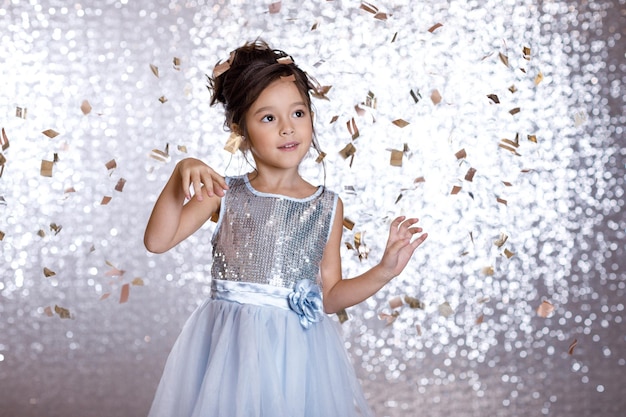 Jolie petite fille souriante en robe argentée et bleue dansant sur fond de confettis avec bokeh argenté. fête d'anniversaire