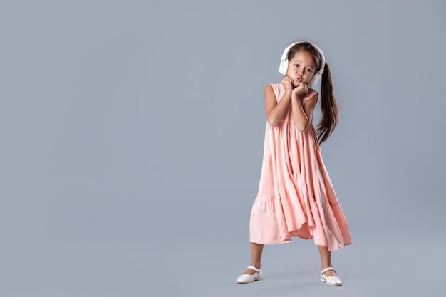 Jolie petite fille souriante en robe rose et casque écoute de la musique et danse sur fond gris.