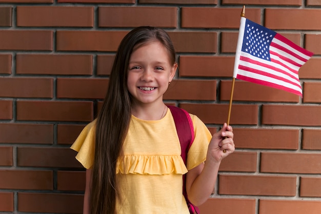 Jolie petite fille souriante et tenant le drapeau américain debout debout contre le mur de briques