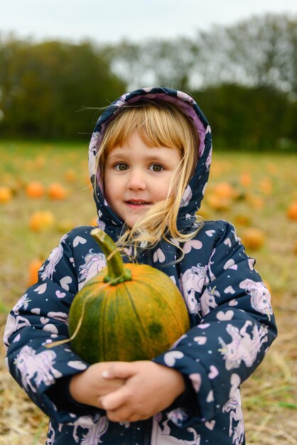 Photo une jolie petite fille souriante en veste d'hiver et en pantalon ramassant une grande citrouille verte et orange