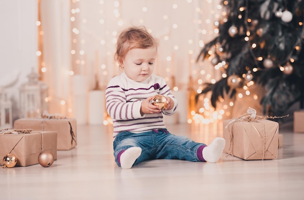 Jolie petite fille tenant une boule de Noël assise sur le sol au-dessus de l'arbre de Noël