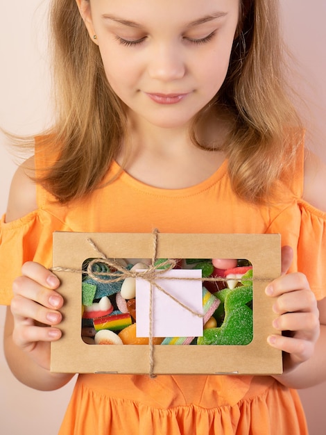 Photo une jolie petite fille tient une boîte pleine de bonbons multicolores dans ses mains