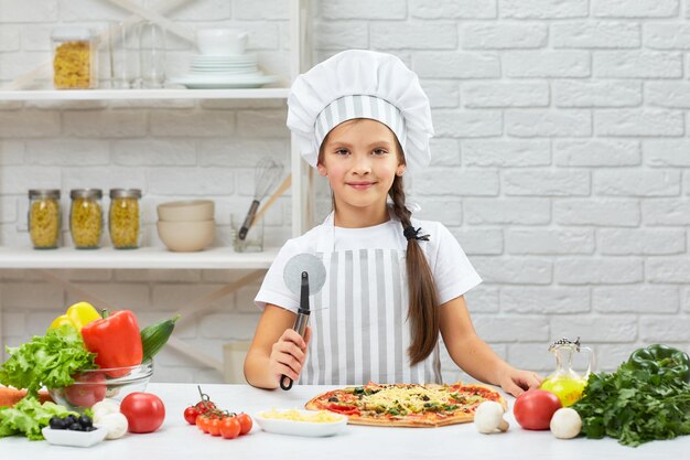 Jolie petite fille en toque et un tablier coupant la pizza