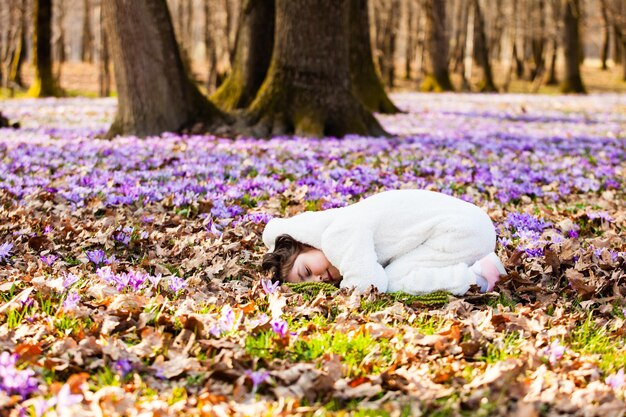 Jolie petite fille en vêtements de nuit moelleux blanc chaud portant sur un sol recouvert de premières fleurs de crocus de printemps prétendant être un petit lapin blanc endormi