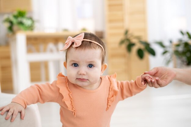 Une jolie petite fille vêtue d'un body rose se tient dans le salon de la maison tenant au support les premiers pas de l'enfant que le bébé apprend à marcher