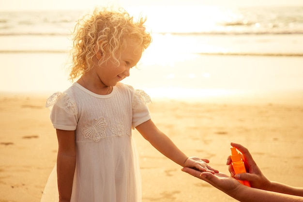 Jolie Petite Fille Vêtue D'une Robe Blanche De Longs Cheveux Blonds Se Relaxant Sur La Plage Près De La Mer, été, Vacances, Concept De Voyage. Souriante Mignonne Petite Fille En Vacances à La Plage Spf Et Crème Solaire.