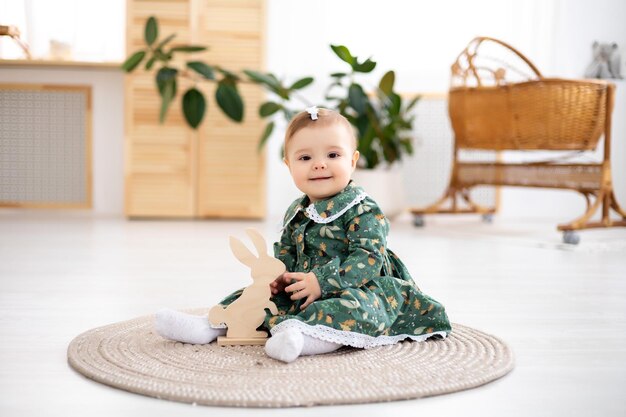 Une jolie petite fille vêtue d'une robe en coton vert est assise sur un tapis dans le salon de la maison en regardant la caméra en souriant jouant avec un lièvre en bois