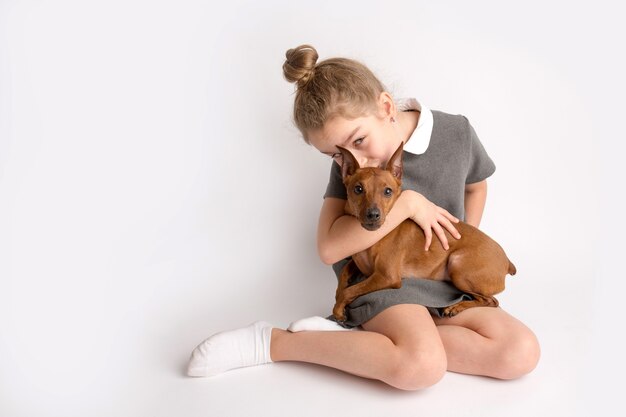 Jolie petite fille vêtue d'une robe d'école stricte avec un pinscher brun nain sur fond blanc, admire une expression heureuse sur son visage. photo de haute qualité