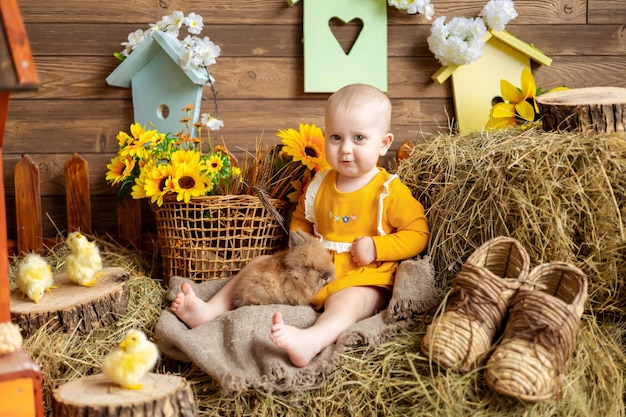 Une jolie petite fille vêtue d'une robe orange est assise sur un foin sur un fond sombre avec des oeufs de Pâques parmi des fleurs avec des lapins et des accessoires de Pâques Célébration de la Grande Pâques Traditions de Pâques