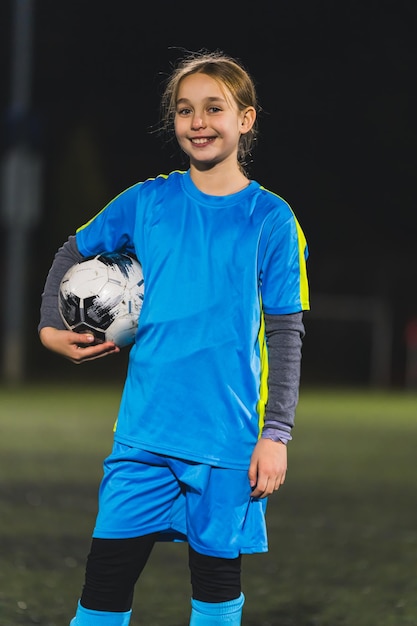 Une jolie petite footballeuse caucasienne dans un uniforme bleu posant avec un ballon de football moyen tiré