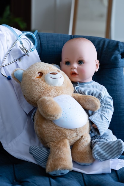 une jolie poupée avec un ours en peluche doux est assise sur un fauteuil bleu à côté d'une blouse médicale