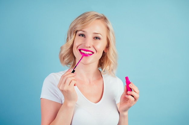 Jolie et souriante femme blonde applique le maquillage de rouge à lèvres rose fuchsia lèvres en studio sur fond bleu