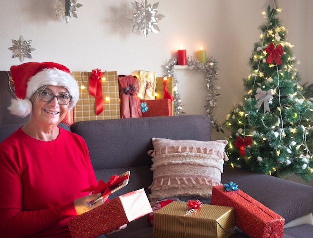 Jolie vieille dame avec un chapeau de Père Noël ouvre son cadeau de Noël en trouvant une nouvelle tablette. Arbre de Noël et cadeaux pour la famille autour d'elle