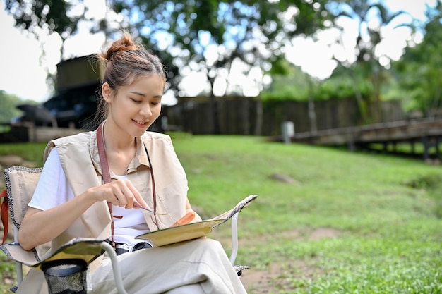 Jolie voyageuse asiatique assise sur la chaise de camping et déjeunant au camping