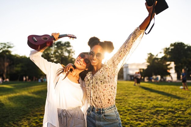 Jolies filles souriantes avec une petite guitare et un appareil photo polaroid levant joyeusement les mains tout en passant du temps ensemble dans le parc