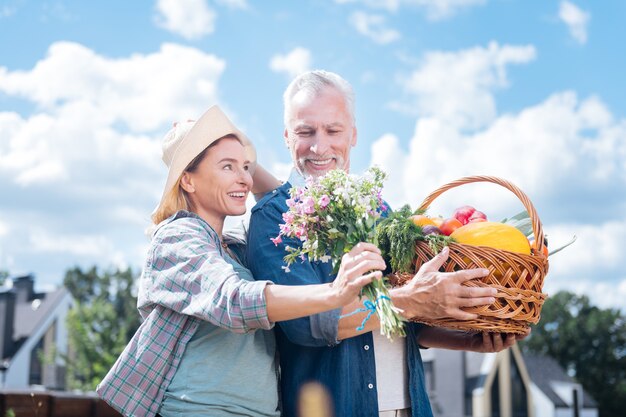 Jolies fleurs. Mari attentionné aux cheveux gris se sentant bien après avoir présenté des fleurs sur le terrain à sa belle femme rayonnante