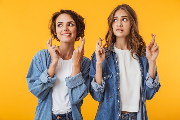 Jolies jeunes femmes amis posant isolé sur mur jaune font espoir s'il vous plaît geste.