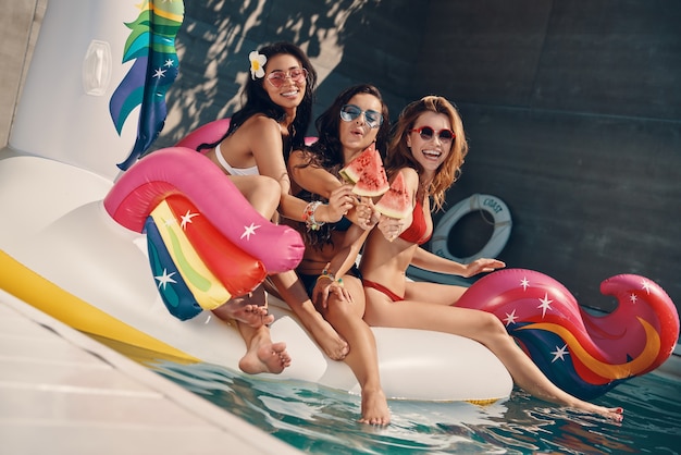 Jolies jeunes femmes en maillot de bain souriant et mangeant de la pastèque tout en flottant sur une licorne gonflable dans une piscine à l'extérieur