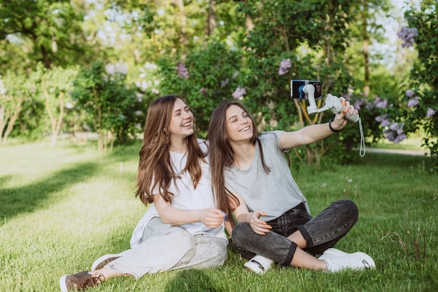 De jolies jeunes femmes souriantes et blogueuses influenceuses filment ou enregistrent des vidéos avec leur smartphone sur un stabilisateur, dans un parc verdoyant et ensoleillé à l'extérieur. Notion de blog. Mise au point sélective douce.