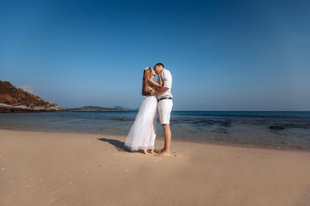 Jolies jeunes mariées mignonnes en vêtements blancs s'embrassant sur une plage de sable le jour de leur mariage au fond de la mer et du ciel. Notion de romantisme.