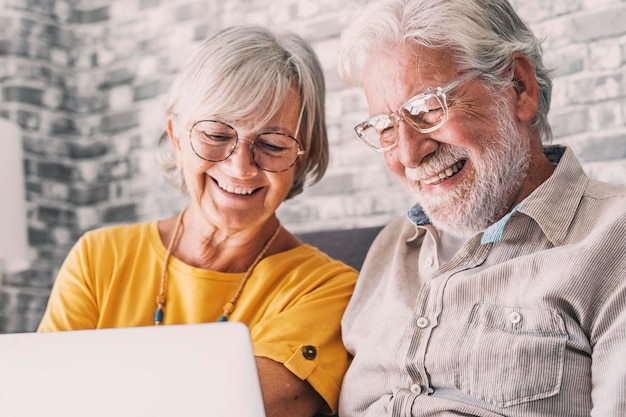 Jolies personnes âgées des années 70 couple aux cheveux gris se reposant sur le canapé dans le salon tenir sur l'ordinateur portable regarder un film souriant profiter du temps libre génération plus âgée et la technologie sans fil moderne concepts d'utilisateurs avancés