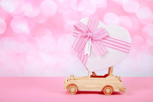 Jouet de voiture avec un cadeau et un arc en forme de coeur sur fond rose avec bokeh