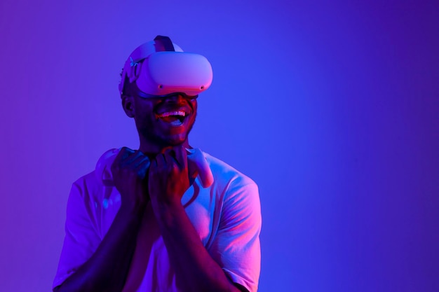 un joueur afro-américain portant des lunettes de réalité virtuelle avec des joysticks regarde ailleurs dans un éclairage au néon