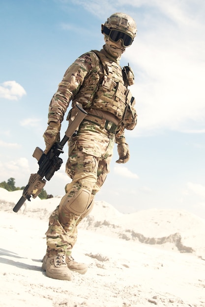 Joueur d'airsoft, participant à des jeux militaires en uniforme de camouflage d'infanterie de l'armée américaine, masque tactique, casque et lunettes, debout dans une zone désertique avec un fusil de service de combat ou une réplique de carabine dans les mains