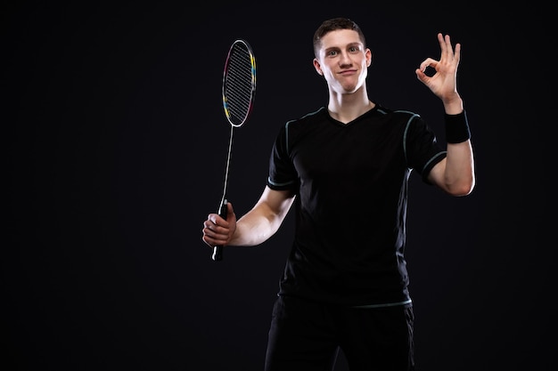 Joueur de badminton en vêtements de sport avec raquette et volant sur fond noir Portrait de sport émotionnel avec homme qui montre ok et bonne chance