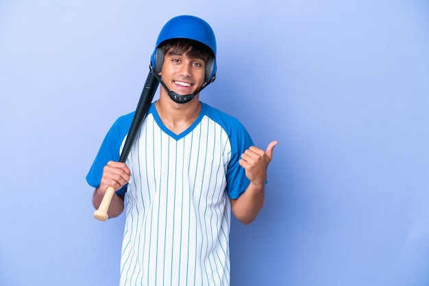 Joueur de baseball homme caucasien avec casque et batte isolé sur fond bleu pointant vers le côté pour présenter un produit