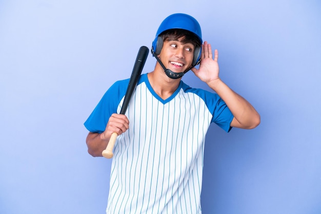 Joueur de baseball homme caucasien avec casque et chauve-souris isolé sur fond bleu écoutant quelque chose en mettant la main sur l'oreille