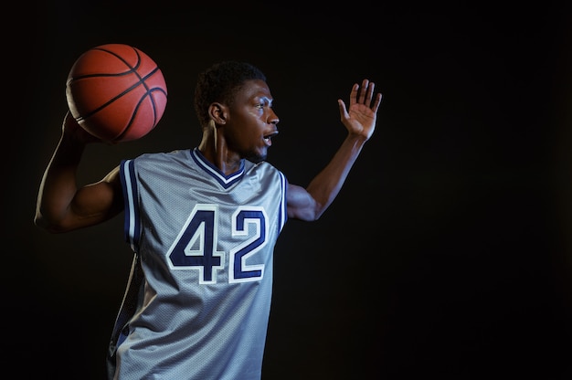 Joueur de basket agressif pose avec ballon en studio, fond noir. Baller professionnel en vêtements de sport jouant à un jeu de sport, grand sportif