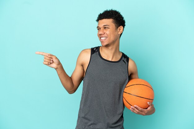 Joueur de basket-ball homme afro-américain isolé sur fond bleu, pointant le doigt sur le côté et présentant un produit