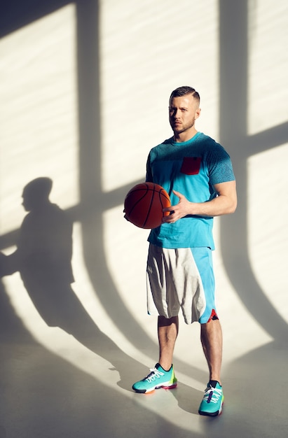 Joueur de basket-ball de jeune homme athlétique tenant le ballon et debout près du mur avec des ombres de la fenêtre