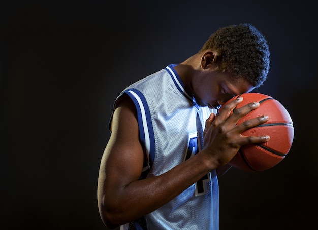 Joueur de basket ciblé pose avec ballon en studio, fond noir. Baller professionnel en vêtements de sport jouant à un jeu de sport, grand sportif