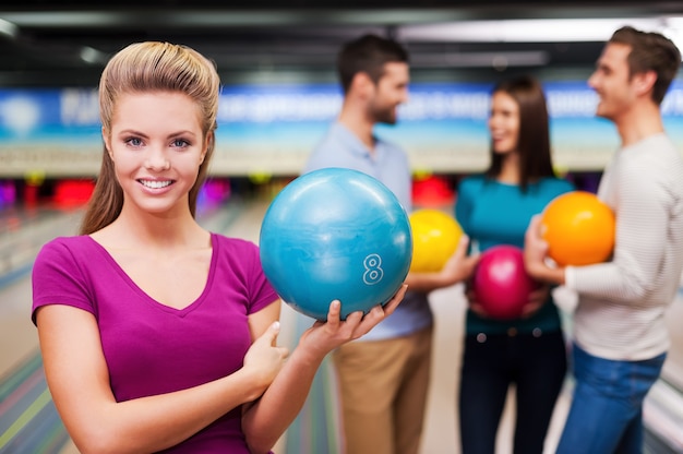 Joueur beau et confiant. Belles jeunes femmes tenant une boule de bowling tandis que trois personnes communiquent contre des pistes de bowling