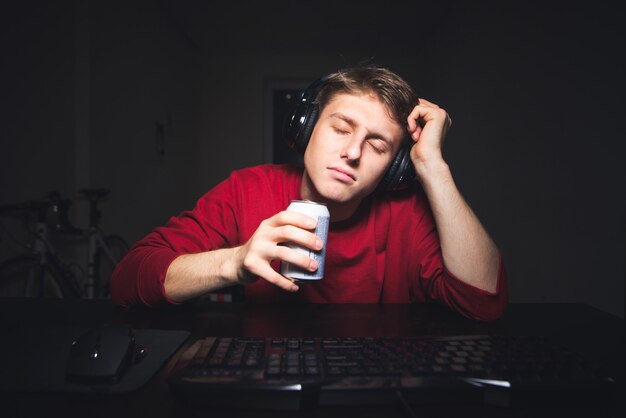 Joueur endormi avec un casque assis à la table avec une canette de boisson à la main et endormi