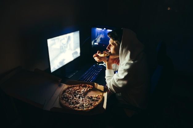 Le joueur est assis à la maison devant l'ordinateur la nuit utilise l'ordinateur et mange de la pizza prête à l'emploi