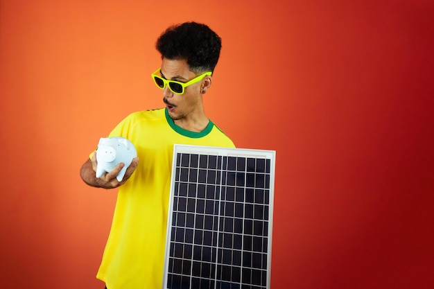 Joueur de football Black Man Celebrating Holding Solar Photovoltaic Panel et Pig Coin isolé sur fond orange