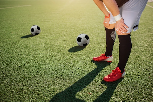 Joueur de football se tenir sur la pelouse verte et tenir les mains sur les genoux. Le soleil brille dehors. Deux ballons de football allongés sur la pelouse plus loin.