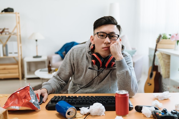 Joueur de gars ennuyé jouant à des jeux vidéo en ligne sur ordinateur tout en étant assis près d'une table en désordre à la maison. homme asiatique paresseux avec des casques malheureux dans une chambre sale. jeune homme à l'intérieur reste à l'intérieur par une chaude journée d'été