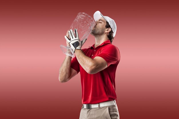 Joueur de golf dans une chemise rouge célébrant avec un trophée de verre dans ses mains, sur fond rouge.