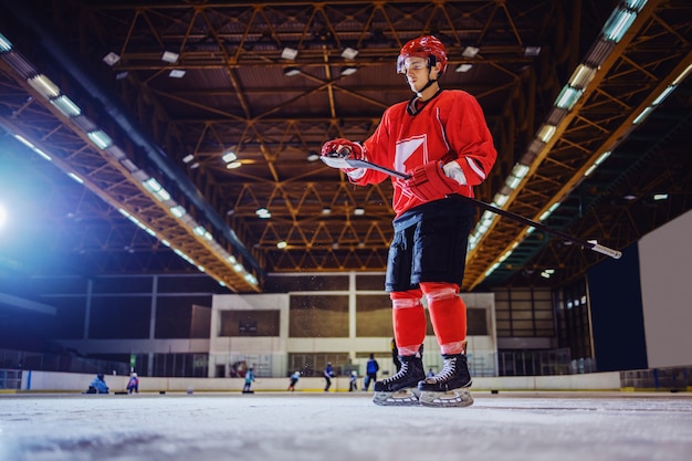 joueur de hockey caucasien debout sur la glace et enlever la glace du bâton. Intérieur du hall. Sports d'hiver.