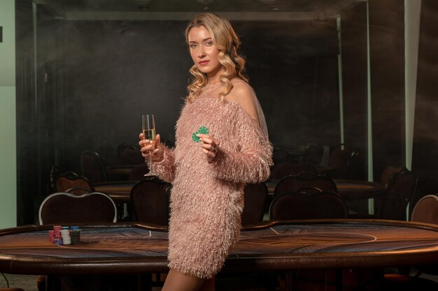 Joueur de poker féminin confiant avec un verre de champagne et des jetons debout près de la table de jeu