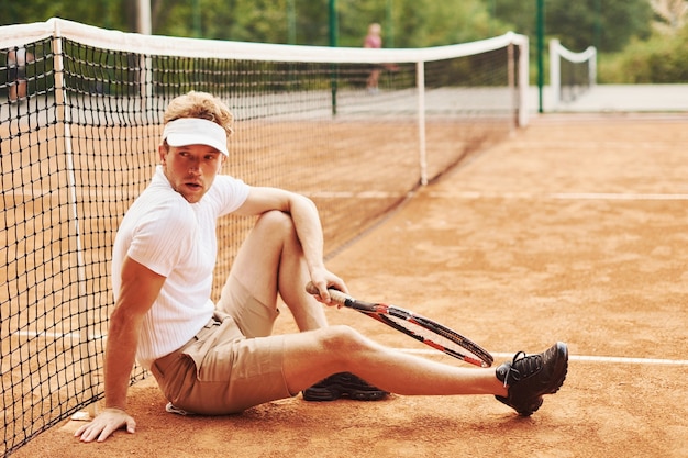 Un joueur de tennis fatigué en tenue sportive est sur le terrain à l'extérieur, appuyé sur le filet.