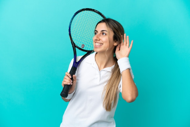 Joueur de tennis de jeune femme isolé sur fond bleu écoutant quelque chose en mettant la main sur l'oreille