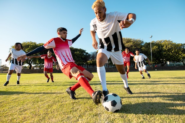 Des joueurs multiraciaux courent et donnent des coups de pied au ballon de football pendant un match sur le terrain de jeu contre un ciel dégagé.