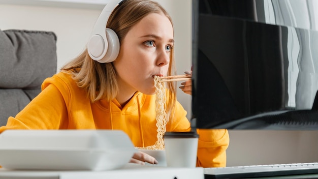 Une joueuse affamée mange des nouilles Plat chinois à l'intérieur de la maison à l'aide d'un ordinateur de bureau pendant un jeu vidéo en streaming