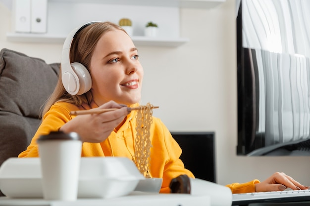 Une joueuse heureuse mange des nouilles avec des baguettes Plat chinois à l'intérieur de la maison à l'aide d'un ordinateur de bureau pendant le jeu vidéo en streaming. Femme adolescente travaillant passionnément la programmation.